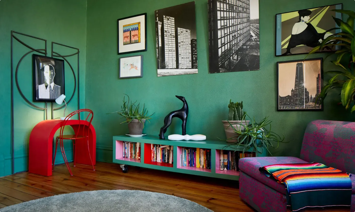 Cozy living room with green walls, wooden floor, red curved desk with matching chair, low green bookshelf with plants and black dog sculpture, colorful blanket on purple patterned sofa, and framed art on walls.